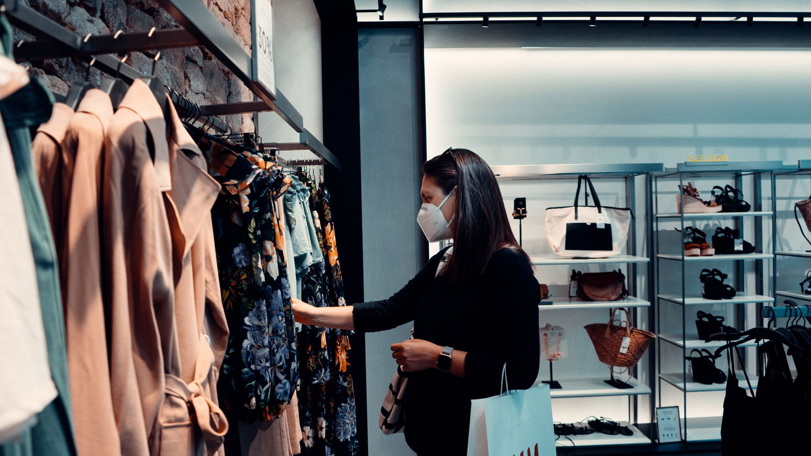 A person with medium toned skin and dark straight hair wearing a white mask shopping, examining a jacket hanging on a rack instead of a consignment store